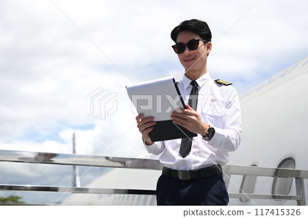 Pilot wearing sunglasses reading paper while standing beside an airplane with cloudy sky on background Pilot wearing sunglasses reading paper while standing beside an airplane with cloudy sky on background 117415326