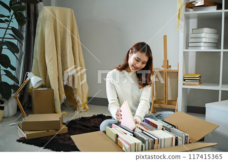 Pretty young asian woman sitting near carton box, packing a pile of books into a box for moving 117415365