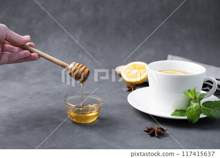 Honey flows from a wooden stick into a glass bowl close-up on a dark background with a cup of tea Honey flows from a wooden stick into a glass bowl close-up on a dark background with a cup of tea 117415637