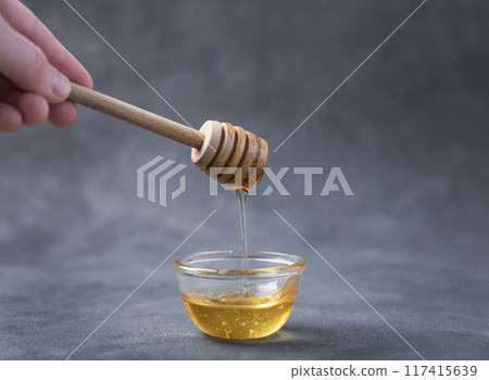 Honey flows from a wooden stick into a glass bowl on a dark background close-up. Honey flows from a wooden stick into a glass bowl on a dark background close-up. 117415639