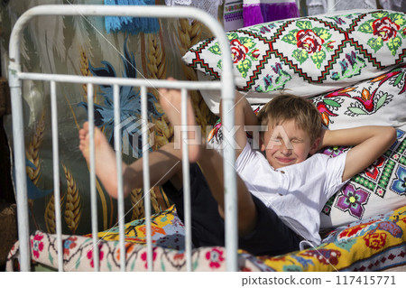 Child Russian or Ukrainian on an old ethnic bed with pillows and bedspreads embroidered with a Slavic pattern. Belarusian boy. 117415771