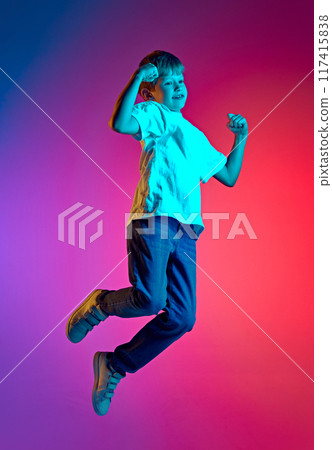 Triumphant young boy jumping clenched fists of joy because holidays start in neon light against gradient blue-pink studio background. Triumphant young boy jumping clenched fists of joy because holidays start in neon light against gradient blue-pink studio background. 117415838