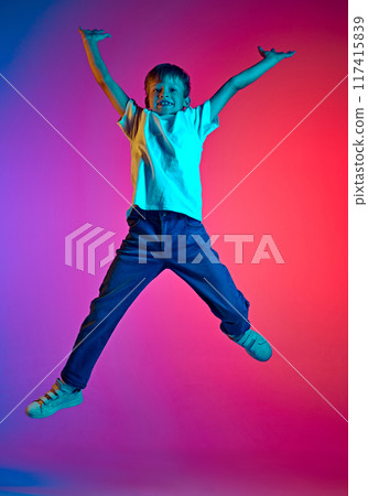 Boy, preteen jumping, raising hands of joy and happiness in neon light against gradient blue-pink studio background. Boy, preteen jumping, raising hands of joy and happiness in neon light against gradient blue-pink studio background. 117415839
