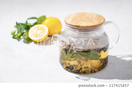 Glass teapot of herbal tea with ginger and mint on a light background with fresh lemon. Glass teapot of herbal tea with ginger and mint on a light background with fresh lemon. 117415857