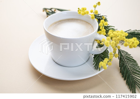 A spring bouquet with yellow mimosa flowers and a cup of coffee cappuccino on a beige background. A spring bouquet with yellow mimosa flowers and a cup of coffee cappuccino on a beige background. 117415902