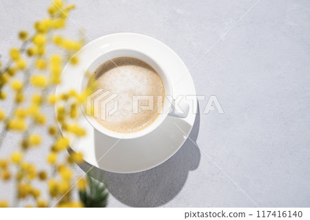 Cappuccino or latte with milk foam in a white cup on a light background with yellow mimosa flowers. 117416140