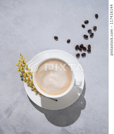 A cup of coffee cappuccino with spring flower mimosa on a light background with coffee beans. A cup of coffee cappuccino with spring flower mimosa on a light background with coffee beans. 117416144