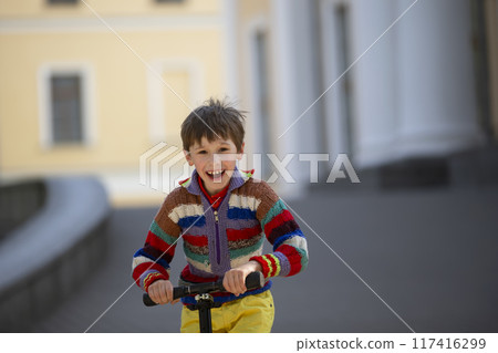 The child is holding the wheel of the scooter. Cheerful boy on a walk in the street. The child is holding the wheel of the scooter. Cheerful boy on a walk in the street. 117416299