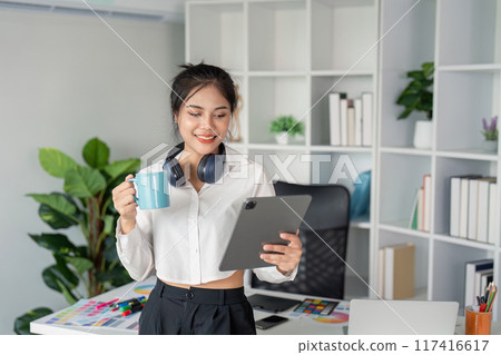 Young Graphic Designer Working in Modern Office with Tablet and Coffee, Surrounded by Creative Tools and Plants Young Graphic Designer Working in Modern Office with Tablet and Coffee, Surrounded by Creative Tools and Plants 117416617