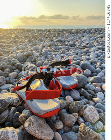 A pair of red and black sandals rest on a pebble beach 117416845