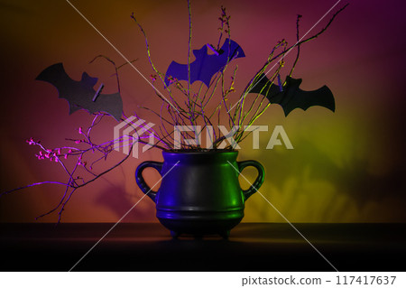 Mysterious black cauldron with dry plants and cutout bats in spotlight on dark neon background. Represents Halloween, crafts, magic, and fantasy concepts. 117417637