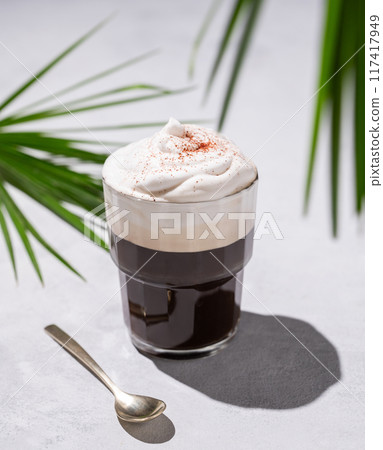 Iced coffee in a glass with foam on a light background with palm leaves, spoon and morning shadows. Iced coffee in a glass with foam on a light background with palm leaves, spoon and morning shadows. 117417949