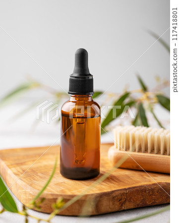 Nail oil or serum in a bottle on a wooden board with a brush on a gray background with green leaves. Nail oil or serum in a bottle on a wooden board with a brush on a gray background with green leaves. 117418801