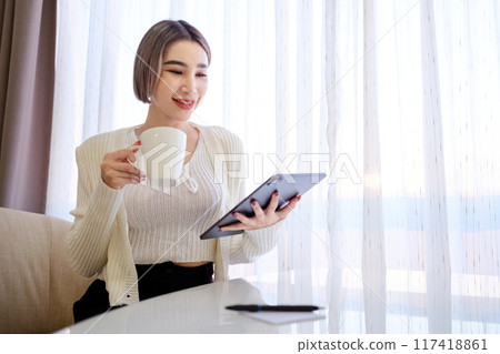 A woman is sitting at a table with a tablet and a cup of coffee A woman is sitting at a table with a tablet and a cup of coffee 117418861