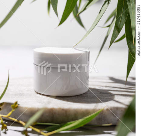 Face and body cream in a white jar on a marble board on a light background with leaves  117419085
