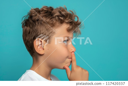 A thoughtful young boy in a white shirt gestures for silence, his eyes full of curiosity. The background's stark blue sets a contrasting stage for his light-hearted secret. 117420337