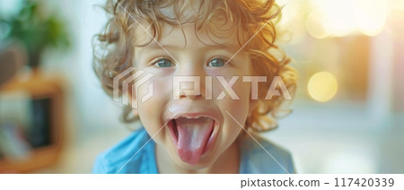 A young boy in a blue shirt exudes happiness with his tongue out, illuminated by a bright and airy indoor light. His curly locks enhance the cheerful scene. 117420339