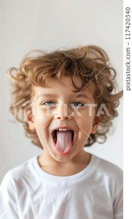A bubbly child with a wide smile and tongue out stands against a simple background, his joy uncontained. His tousled curls frame a face full of life. 117420340
