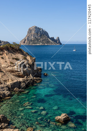 Vertical photo of Es Vedra island in Ibiza with a catamaran on clear blue waters, rocky coast 117420445