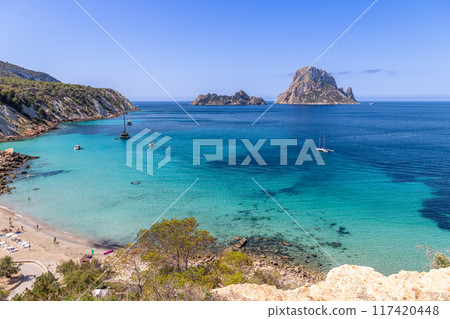 Cala Hort beach in Ibiza, with anchored boats, stunning emerald waters, rugged cliffs 117420448