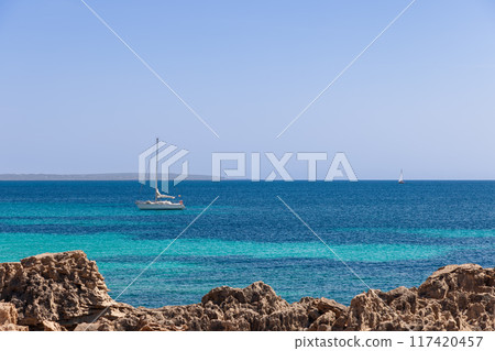 The rugged coast of Ibiza meets pristine, calm waters under an empty sky, with a distant sailboat The rugged coast of Ibiza meets pristine, calm waters under an empty sky, with a distant sailboat 117420457