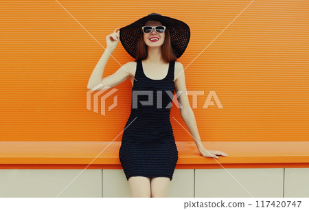 Portrait of beautiful young woman wearing a black summer hat and dress on orange background Portrait of beautiful young woman wearing a black summer hat and dress on orange background 117420747