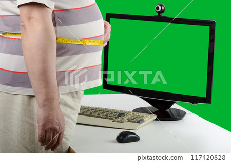 Overweight Man Prepares for Virtual Health Check-Up, Measuring Waist in Front of Green-Screen Monitor, Signifying Telemedicine and Dietary Guidance. 117420828