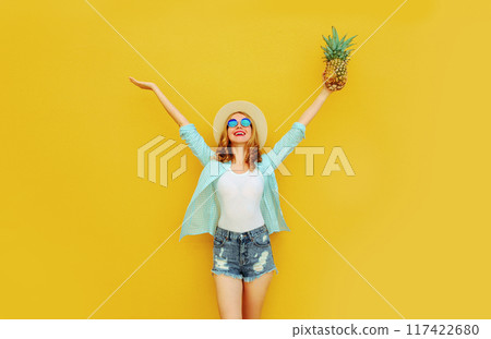 Happy cheerful woman raises her hands up with pineapple have fun in summer hat on yellow background 117422680
