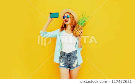 Happy smiling woman with pineapple taking selfie with phone in summer hat on color yellow background 117422681