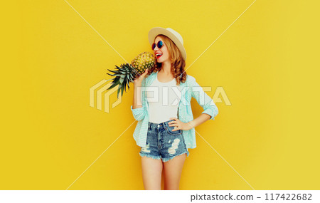 Happy woman with pineapple having fun in summer straw hat, shorts on colorful yellow wall background 117422682