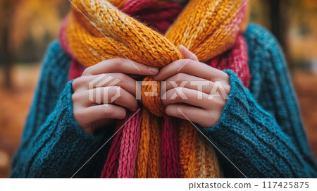 Hands tying a cozy knitted scarf in warm autumn colors with a fall landscape in blurred background 117425875