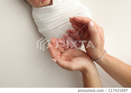 The palms of the parents. A father and mother hold the feet of a newborn child in a white blanket on a white background.. The feet of a newborn in the hands of parents. Photo of foot, heels and toes 117425897