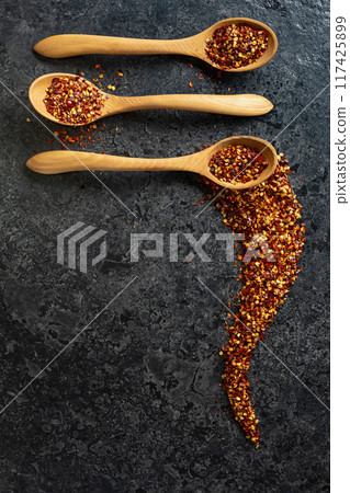 Flakes of red chili pepper in wooden spoons on a black stone background. Flakes of red chili pepper in wooden spoons on a black stone background. 117425899