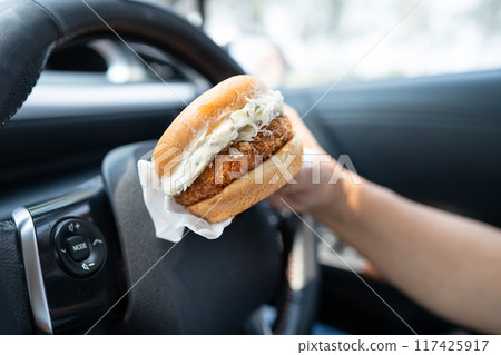 Asian lady holding hamburger to eat in car, dangerous and risk an accident. Asian lady holding hamburger to eat in car, dangerous and risk an accident. 117425917