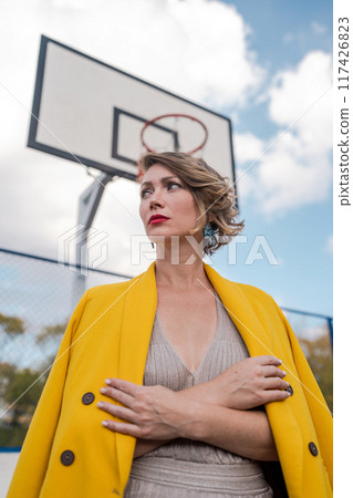 Woman in evening dress and yellow jacket poses against a basketball hoop like a halo. Glamorous lady on basketball court. Fashion, sport concept. Woman in evening dress and yellow jacket poses against a basketball hoop like a halo. Glamorous lady on basketball court. Fashion, sport concept. 117426823