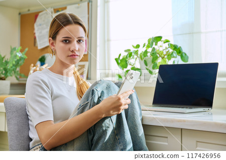 Portrait of smiling teenage girl student sitting at home at desk, holding smartphone Portrait of smiling teenage girl student sitting at home at desk, holding smartphone 117426956