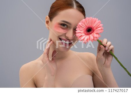 Happy young red haired woman with gerbera on face wearing moisturizing patches on skin 117427017