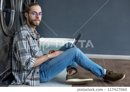 Handsome man accessing internet on laptop at self-service laundry 117427068