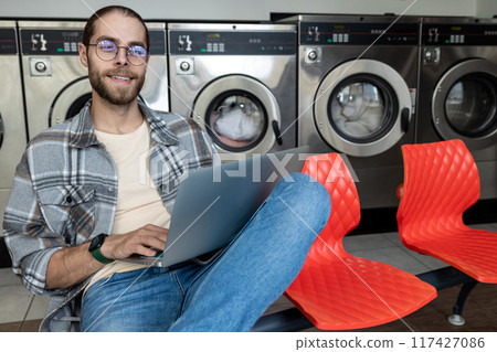 Handsome man accessing internet on laptop at self-service laundry Handsome man accessing internet on laptop at self-service laundry 117427086