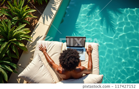 A dark-skinned digital nomad sits near the pool with a laptop. Top view 117427152