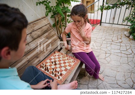 Children engaged in an intense outdoor chess game on a wooden bench, strategizing their next moves in a thoughtful and playful setting. 117427284