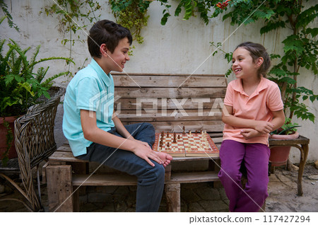 Two cheerful children enjoying a fun game of chess while sitting on a wooden bench in a lush garden setting, showcasing friendship and strategic thinking. 117427294