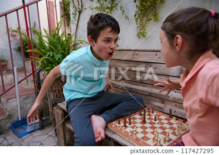 Young boy passionately playing chess with a girl, showing competitive spirit in a friendly backyard atmosphere surrounded by greenery 117427295