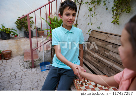 Two children shaking hands after a chess game outdoors, expressing sportsmanship and friendship in a garden setting with rustic wooden bench and green plants around them. Two children shaking hands after a chess game outdoors, expressing sportsmanship and friendship in a garden setting with rustic wooden bench and green plants around them. 117427297