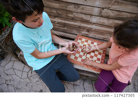 Children sitting on a bench play a board game using a wooden chess set, enjoying a peaceful and leisurely afternoon outdoors together. 117427305