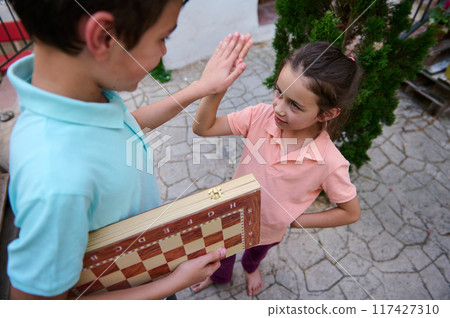 Children enjoying an outdoor high five after a friendly game of chess, showcasing teamwork, camaraderie, and the fun of strategic play in a casual setting. 117427310