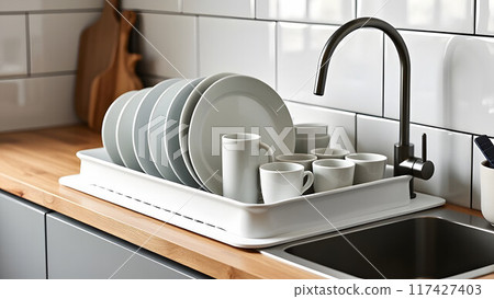 Dish rack holds many dishes and cups against wooden countertop, white wall tiles, sink and faucet. Budget and lightweight antimicrobial dish drainer with drain board at modern scandinavian kitchen 117427403