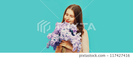 Portrait of beautiful woman enjoying smell of fresh bouquet lilac flowers on blue background 117427416