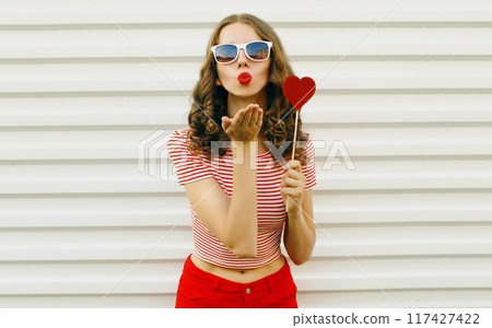 Portrait of beautiful young woman with red heart shaped sunglasses blowing kiss on white background 117427422