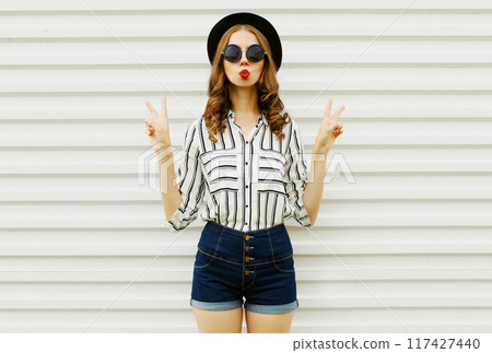 Attractive young woman blowing kiss in black round hat, shorts, striped shirt on white background 117427440